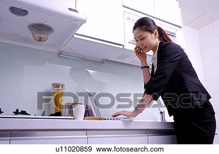 Stock Photo - Young woman on the phone at kitchen counter using laptop computer. Fotosearch