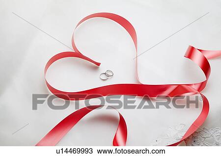 Stock Image - Red ribbon in shape of heart, two wedding rings within heart. Fotosearch