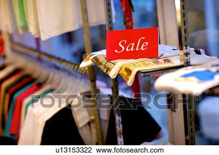 Stock Image - Red and white 'Sale' sign in a clothing store, on top of several T-shirts. Fotosearch
