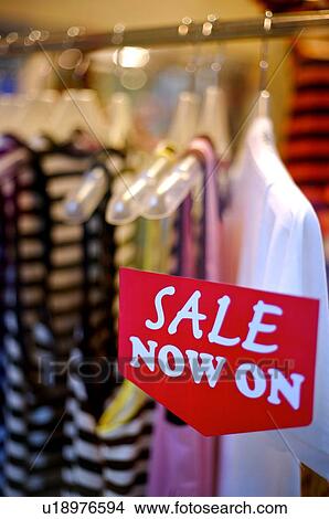 Picture - Red and white 'Sale' sign on the window of a clothing store, in front of a rack of clothes. Fotosearch