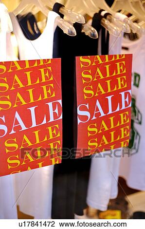 Stock Image - Red 'Sale' posters on the window of a clothing store, with a row of tank tops in the display window. Fotosearch