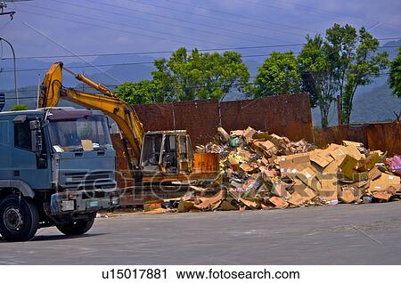 Stock Image - Excavator and truck next to a pile of cardboard boxes. Fotosearch