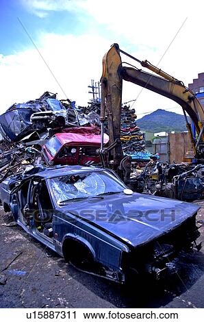 Stock Image - Excavator digging at piles of compressed cars in a junkyard. Fotosearch