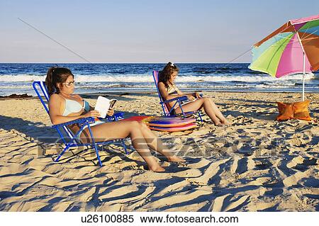 Stock Photography - View of teenager (12 and 16 years old) reading on beach and beach umbrella at sunset, New Jersy, USA. Fotosearch
