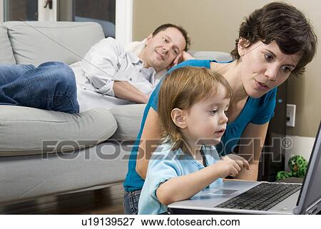 Stock Photo - Woman and child using laptop on coffee table, man behind on sofa, Winnipeg, Canada. Fotosearch