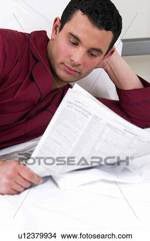 Picture - Hispanic man reading newspaper while lying in bed. Fotosearch