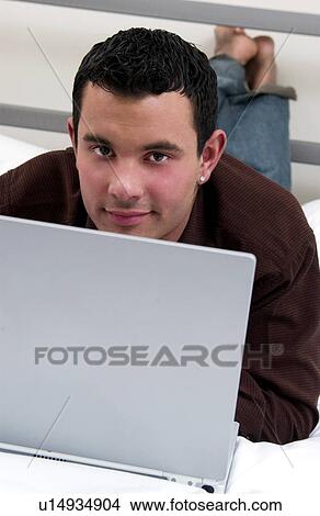 Picture - Hispanic man working on laptop computer while lying on bed. Fotosearch