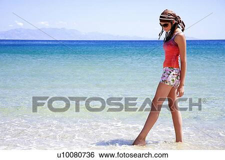 Stock Photograph - Young woman on beach with her feet in water. Fotosearch