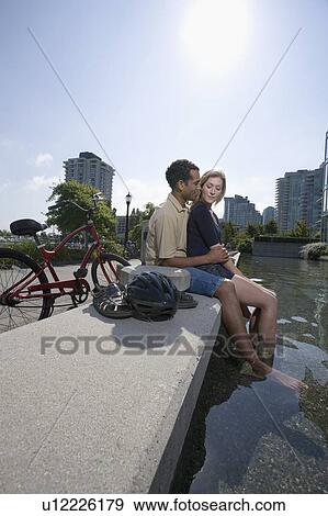 Stock Photo - Young Couple rest from riding bikes to cuddle at a fountain, side view, Vancouver, British Columbia. Fotosearch