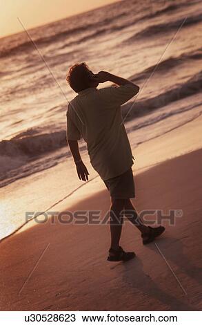 Stock Image - Man talking on cell phone while walking on beach. Fotosearch