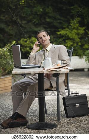 Stock Image - Businessman in garden cafe with cell phone and laptop. Fotosearch