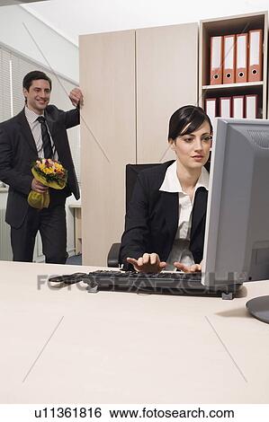 Stock Photograph - Male office worker about to surprise female colleague with flowers. Fotosearch