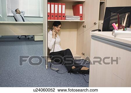 Stock Image - Office worker on the phone hiding from manager. Fotosearch