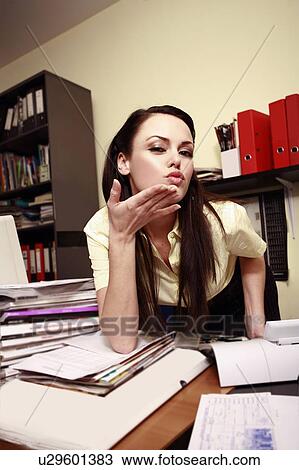 Stock Image - Female office worker at cluttered desk blowing a kiss. Fotosearch