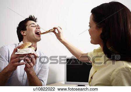 Stock Image - Office worker feeding her colleague at desk. Fotosearch