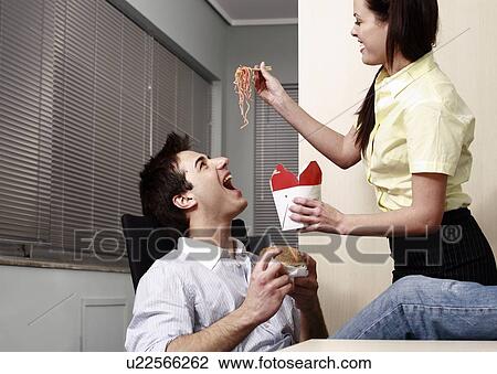 Stock Image - Office worker feeding her colleague at desk. Fotosearch