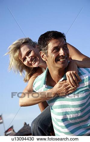 Stock Image - Mature adult couple piggyback on beach. Fotosearch