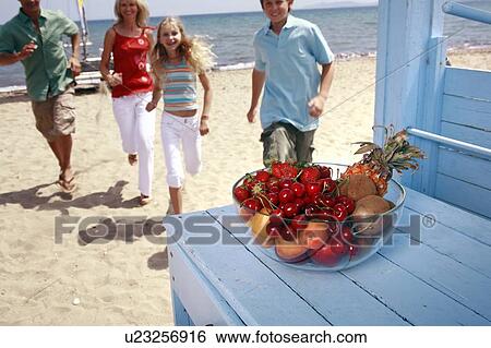 Stock Photograph - Bowl of fruit and parents with children on beach. Fotosearch