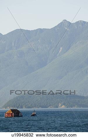 Stock Image - Tug Boat pulling logs through the ocean with mountains in the background, Howe Sound, BC. Fotosearch
