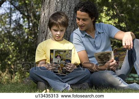 Stock Image - Father and son beside tree reading comic books, Assiniboine Park, Winnipeg, Canada. Fotosearch