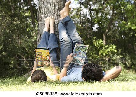 Stock Photography - Father and son lying on backs with feet resting on tree, reading comic books, Assiniboine Park, Winnipeg, Canada. Fotosearch