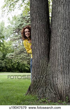 Stock Photo - Young woman peeks out from behind tree, Winnipeg, Manitoba, Canada. Fotosearch