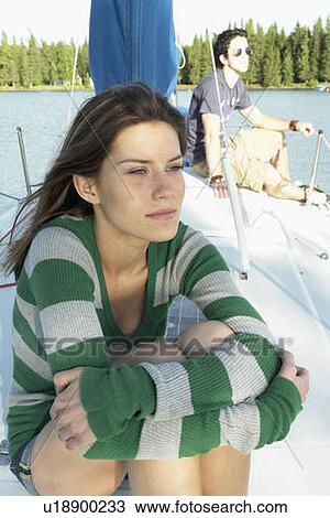 Stock Image - Young woman in foreground, young man in background, on sailboat, Clear Lake, Riding Mountain National Park, Manitoba, Canada. Fotosearch