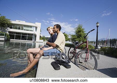 Stock Image - Young Couple rest from riding bikes at a fountain, side view, Vancouver, British Columbia. Fotosearch