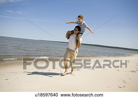 Stock Image - Man carries boy (7) on his shoulders as boy holds arms out, Grand Beach Provincial Park, Manitoba, Canada. Fotosearch