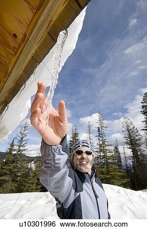 Stock Photograph - A woman grabs an icicle from the roof of a winter cabin, Silver Star ski resort near Vernon, BC, Canada. Fotosearch