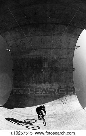 Stock Photography - Black and white mountain biker dwarfed by concrete pipe; Canada, Alberta, Calgary, Shaw Millenium Park. Fotosearch
