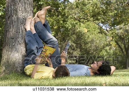 Stock Photo - Father and son lying on backs with feet resting on tree, reading comic books, Assiniboine Park, Winnipeg, Canada. Fotosearch