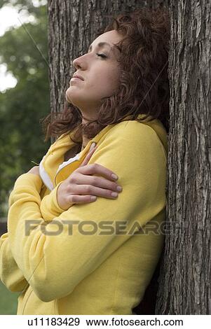 Stock Photo - Side view of woman with eyes closed leans against trunk of tree, Munson Park, Winnipeg, Canada. Fotosearch