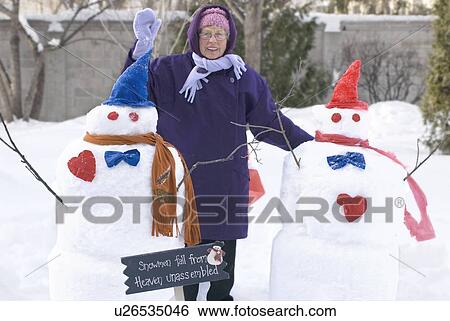 Stock Photograph - Senior woman waving beside snowman that she made, Winnipeg, Canada. Fotosearch