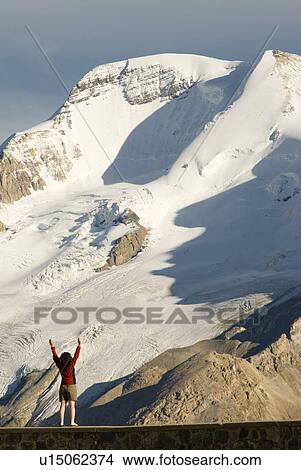 Picture - Woman (20-25) standing on a rock wall with hands raised in front of a large glacier clad peak, Columbia Icefields, Jasper National Park, Alberta, Canada. Fotosearch