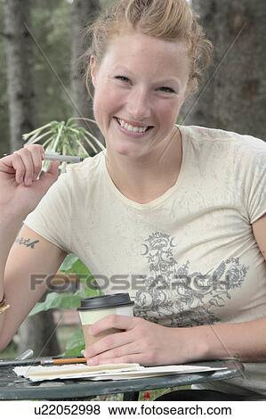 Stock Photo - Woman seated at outdoor restaurant, corn-based compostable coffee cup on table, Wasagaming, Riding Mountain National Park, Manitoba, Canada. Fotosearch