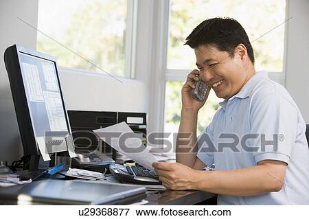 Stock Photo - Man in home office with computer and paperwork on telephone smiling. Fotosearch