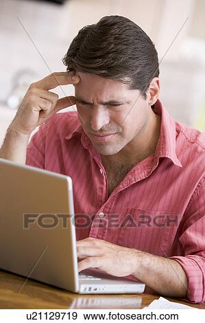 Stock Photo - Man in kitchen using laptop frowning. Fotosearch
