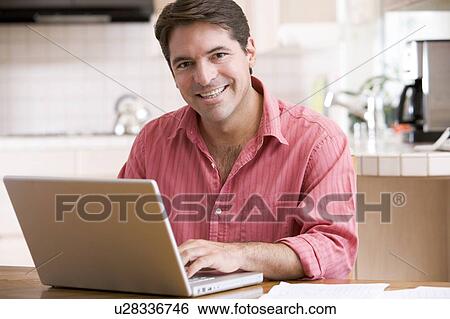 Stock Photograph - Man in kitchen using laptop smiling. Fotosearch
