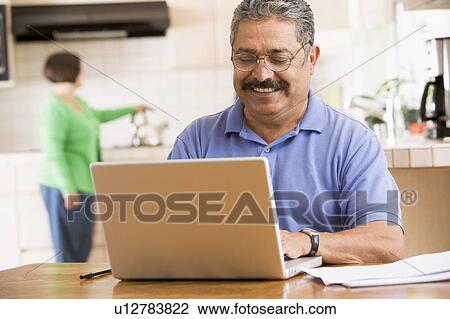 Stock Image - Man in kitchen with laptop smiling with woman in background. Fotosearch