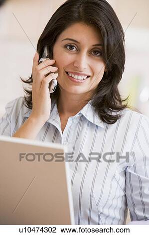 Stock Image - Woman in kitchen with laptop and cellular phone smiling. Fotosearch