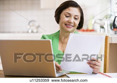 Stock Photo - Woman in kitchen with laptop and paperwork smiling. Fotosearch