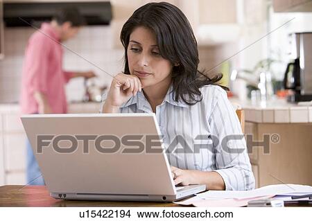 Woman in kitchen with paperwork using laptop with man in background View Large Photo Image Picture - Woman in kitchen with paperwork using laptop with man in background. Fotosearch