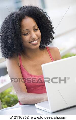 Stock Image - Woman sitting at table outdoors with laptop smiling (high key). Fotosearch