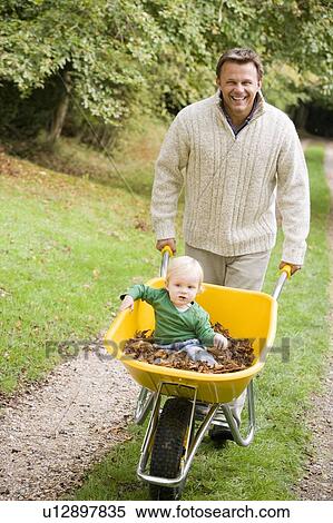 Man walking on path outdoors pushing baby in wheelbarrow and smiling (selective focus) View Large Photo Image Stock Photography - Man walking on path outdoors pushing baby in wheelbarrow and smiling (selective focus). Fotosearch