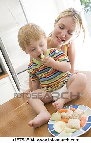 Picture - Mother and baby in kitchen eating fruit and vegetables. Fotosearch