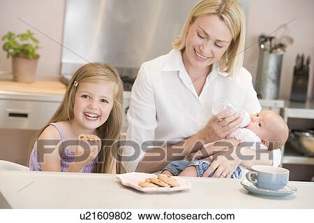 Mother feeding baby in kitchen with daughter eating cookies and smiling View Large Photo Image Stock Image - Mother feeding baby in kitchen with daughter eating cookies and smiling. Fotosearch