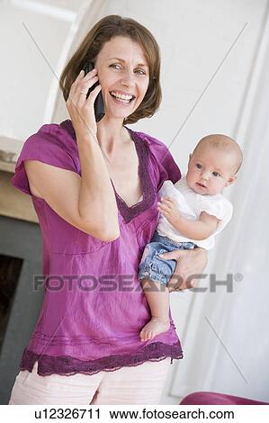 Stock Image - Mother in living room using telephone holding baby smiling. Fotosearch