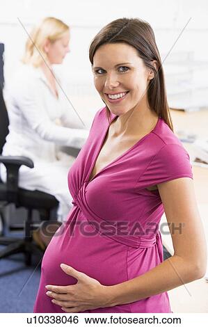 Pregnant woman at work holding belly smiling with coworker in background View Large Photo Image Stock Photograph - Pregnant woman at work holding belly smiling with coworker in background. Fotosearch