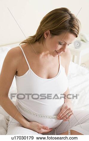 Stock Photo - Pregnant woman in bedroom measuring belly smiling. Fotosearch
